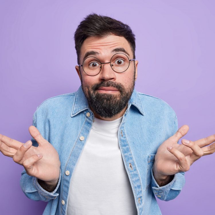 Hesitant indecisive bearded European man spreads palms feels clueless and uncertain canot make choice wears round eyeglasses denim shirt poses against purple background smirks looks doubtful.