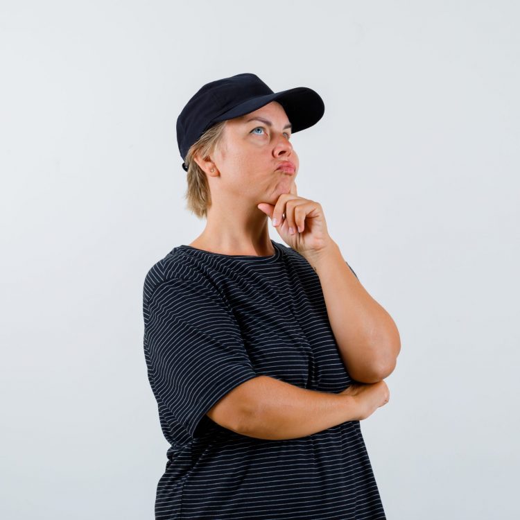 blonde woman leaning chin on fist, thinking about something in black t-shirt, cap and looking pensive , front view.