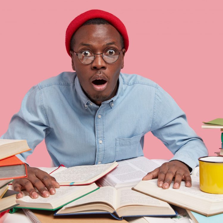 Stupefied workaholic in red headgear, spectacles and formal shirt, stares at camera with frightened facial expression, keeps hands on textbooks, has dark skin, cant believe in negative news.